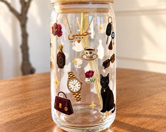 May include: A clear glass tumbler with a bamboo lid and a curved yellow glass straw. The exterior features colourful decals of a black cat, a gold wristwatch, a maroon handbag, a teacup, sunglasses, a perfume bottle, and Arabic text.