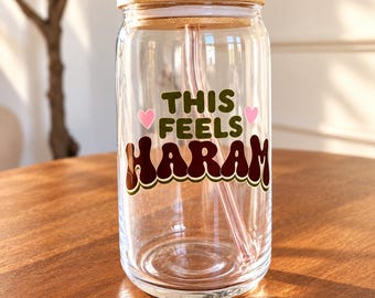 May include: A clear glass tumbler with a bamboo lid and a curved glass straw. The front features the text "THIS FEELS HARAM" in dark brown and green retro-style lettering, accented by two small pink hearts on either side of the word "FEELS."
