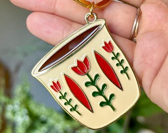 May include: A gold-toned metal keyring featuring a cream-coloured, cup-shaped enamel charm. The charm is decorated with a repeating pattern of red tulips and green stems, with a dark red interior rim. The keyring includes a circular ring and a clasp.
