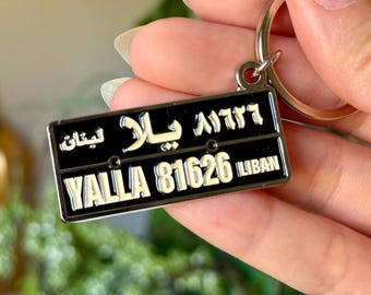 May include: A rectangular black metal keyring featuring a silver-toned border and a silver lobster clasp. The keyring displays the text YALLA 81626 LIBAN in white, along with Arabic script characters in white across the top section of the plate.