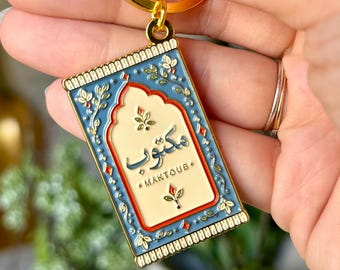May include: A gold-toned metal keyring featuring a rectangular enamel charm shaped like a prayer rug. The charm has a blue border with floral details and a cream-coloured centre with the Arabic text Maktoub and the word MAKTOUB below it.