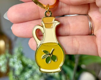 May include: A gold-toned metal keyring featuring an enamel charm shaped like a glass cruet filled with yellow olive oil. The charm displays a small green olive branch graphic on the front and is attached to a circular ring and lobster clasp.