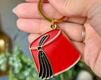 May include: A red enamel keyring shaped like a fez hat with a black tassel detail. The charm features gold-toned metal edges and is attached to a gold-toned split ring and a lobster claw clasp for secure attachment to bags or keys.