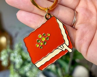 May include: A gold-toned metal keyring featuring a square-shaped enamel charm. The charm depicts a slice of red cake with a white cream layer and green circular toppings, suspended from a circular ring and a lobster claw clasp.