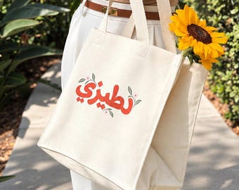 May include: A cream-coloured canvas tote bag featuring red Arabic script text and small green leaf accents on the front. The bag has two sturdy fabric handles and contains a bright yellow sunflower tucked inside the main compartment.