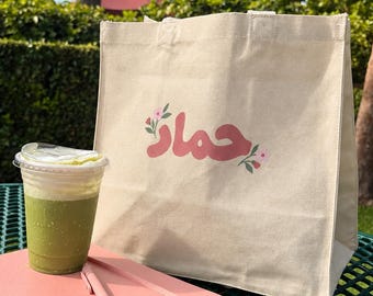 May include: A beige canvas tote bag featuring pink Arabic calligraphy and small floral accents, a pink spiral-bound notebook with a matching pink pen, and a clear plastic cup filled with a green matcha latte topped with white foam.