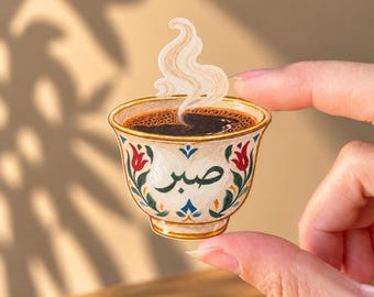 May include: A die-cut sticker shaped like a small coffee cup filled with dark liquid and rising steam. The cup is white with a floral pattern in red, green, and blue, featuring the Arabic word Sabr in black script on the front centre.