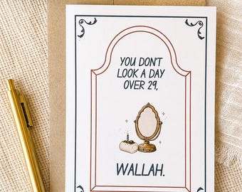 May include: A white greeting card with a dark blue and red border featuring the text "YOU DON'T LOOK A DAY OVER 29, WALLAH." The illustration shows a small cake with a single lit candle next to a gold-coloured standing mirror with sparkles.