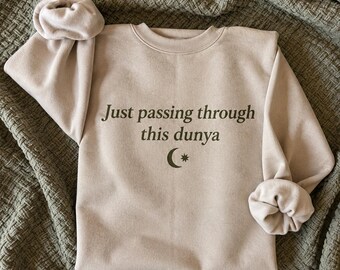 May include: A beige crewneck sweatshirt featuring the dark green text Just passing through this dunya with a crescent moon and star symbol underneath. The garment has long sleeves with gathered cuffs and a ribbed neckline.