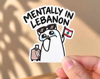 May include: A die-cut sticker featuring a white cartoon character with black sunglasses on its head, crying, holding a small Lebanese flag, and pulling a pink suitcase with a luggage tag labelled Beirut, with the text MENTALLY IN LEBANON above.