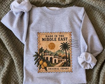 May include: A light grey crewneck sweatshirt featuring a rectangular graphic print designed like a postage stamp. The print displays the text MADE IN THE MIDDLE EAST and ORIGINAL EXPORT, with illustrations of a palm tree, olive branch, and buildings.