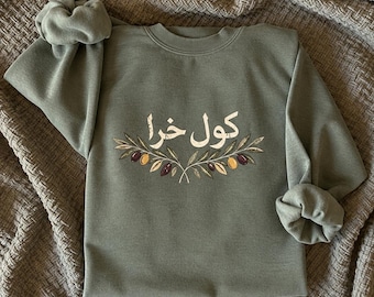May include: An olive green crewneck sweatshirt featuring white Arabic text centred above a graphic illustration of two olive branches with green, black, and yellow olives. The long-sleeved garment has ribbed cuffs and a ribbed neckline.