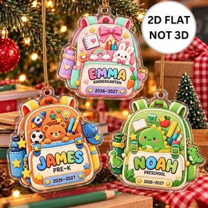 May include: Three 2D flat acrylic ornaments shaped like backpacks in pink, orange, and green. Each features a cartoon character, school supplies, and personalized text including names like Emma, James, and Noah, grade levels, and the year 2026-2027.