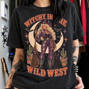 May include: A charcoal gray t-shirt featuring a graphic print of a blonde figure in a purple western outfit and witch hat, holding a broomstick. The design includes a crescent moon, cacti, bats, and a black cat, with the text WITCHY IN THE WILD WEST.