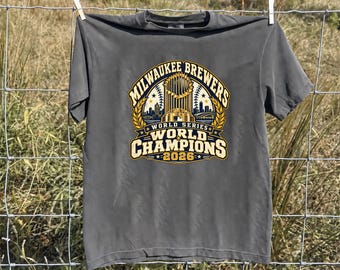 May include: A charcoal gray short-sleeve t-shirt featuring a graphic print that reads Milwaukee Brewers World Series World Champions 2026, centered with an illustration of a trophy, baseball stitching, and a city skyline silhouette.