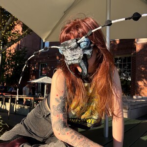 Könnte beinhalten: Eine Maske, die einem Insektenkopf ähnelt, mit großen blauen Augen, strukturierten schwarzen und grauen Oberflächen und langen, dünnen Antennen mit schwarz-weißen Segmenten. Die Maske bedeckt das obere Gesicht und hat markante Mandibeln.