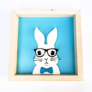 May include: A white bunny with black glasses and a blue bow tie is framed in a light wood frame against a blue background.