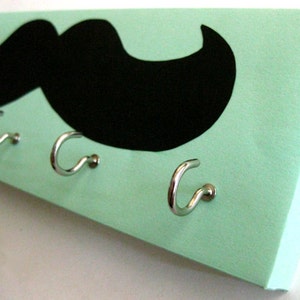 May include: A light green wooden key holder with a black mustache design and five silver hooks.