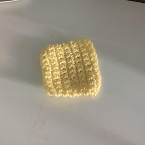 May include: A small, square-shaped crochet piece crafted from pale yellow yarn. The item features a consistent, textured stitch pattern throughout its surface, creating a dense and uniform appearance with slightly raised, rounded edges on all four sides.