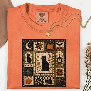 May include: An orange t-shirt with a Comfort Colors label at the neck, featuring a square graphic print of black cats, pumpkins, moons, stars, bats, a spider web, and a potion bottle. The shirt is displayed with a gold star necklace.