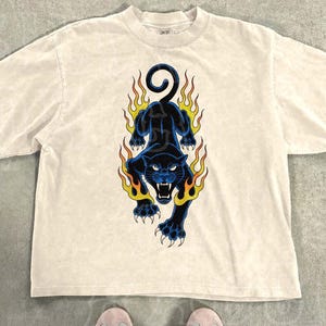 Könnte beinhalten: Ein beiges T-Shirt mit kurzen Ärmeln und einer Grafik eines schwarzen Panthers, der von orangefarbenen und gelben Flammen umgeben ist. Der Text "THE SILLY RACCOON CO" steht oben links und das Wort "natural" unten rechts auf dem Bild.