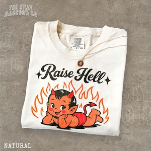 Pode incluir: Uma t-shirt de cor natural com um gráfico de um personagem de diabo vermelho deitado em frente a chamas cor de laranja. O texto "Raise Hell" está impresso a preto acima da personagem. Um colar dourado com um pequeno pendente repousa na peça.