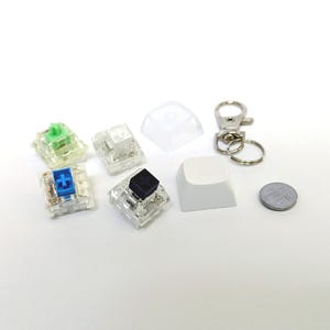 May include: A collection of mechanical keyboard components including four clear switches with green, blue, black, and clear stems, two white keycaps, a silver metal keychain ring, and a small circular gray coin-like object on a white surface.