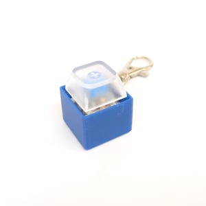 May include: A blue square base holding a single mechanical keyboard switch topped with a translucent white keycap. The switch features a blue stem with a cross-shaped mount. A small silver-toned metal lobster clasp keychain is attached to the side.