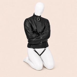 May include: A black leather straightjacket featuring a high collar with a silver-toned metal ring, long sleeves secured across the chest with a vertical strap, and a lower strap attachment connected to a crotch piece with silver-toned hardware.