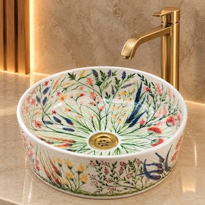 May include: A round ceramic vessel sink featuring a hand-painted botanical pattern of colorful wildflowers, grasses, and foliage in shades of red, blue, yellow, and green. The basin includes a central gold-toned drain and a matching gold faucet.