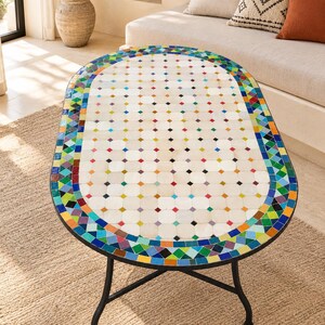 Peut inclure: Une table basse ovale dotée d'une surface en carrelage blanc avec de petits accents carrés multicolores. La table est bordée d'une frise vibrante de carreaux de mosaïque bleus, verts, orange et jaunes sur une base en métal noir à quatre pieds.