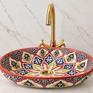 May include: An oval ceramic vessel sink featuring a vibrant, hand-painted floral pattern in red, blue, yellow, and green. The sink includes a central brass drain and is paired with a tall, curved, polished gold-tone faucet with two handles.