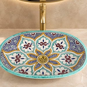 May include: An oval-shaped ceramic vessel sink featuring a vibrant, hand-painted floral pattern in shades of turquoise, deep blue, yellow, and maroon. The basin has a central gold-toned drain and is positioned beneath a tall, brushed gold faucet.
