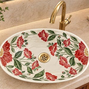 May include: An oval ceramic vessel sink featuring a cream-colored base with a hand-painted floral pattern of red flowers and green leaves. The sink includes a central gold-toned drain and is paired with a matching gold-toned curved faucet.