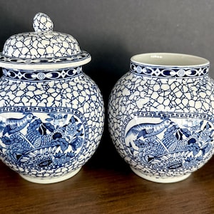 May include: Two round ceramic jars with a white and blue crackle glaze finish. Each jar features a central cartouche containing a detailed illustration of a bird perched in a basket of flowers, framed by a geometric Greek key border pattern.