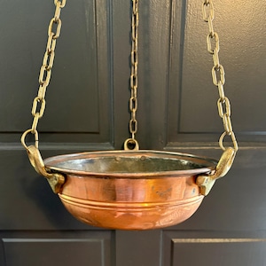 May include: A round copper bowl with a metallic interior, suspended by three brass chains attached to two brass handles on opposite sides. The bowl features a smooth, polished exterior surface and a wide rim, hanging from a central brass ring.