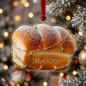 May include: A clear acrylic Christmas ornament shaped like a loaf of bread with a golden brown crust and a dusting of white flour. The name MASON is printed in gold lettering on the front, and the ornament is wrapped in thin gold wire with tiny multicolored LED lights, hanging from a red velvet ribbon.