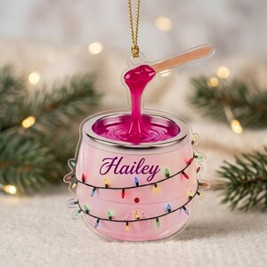 May include: A clear acrylic ornament shaped like a pink wax warmer with a wooden applicator stick dipping into bright pink wax. The warmer features the name Hailey in purple script and is wrapped in a string of colorful Christmas lights.