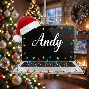May include: A clear acrylic ornament shaped like a laptop computer, featuring a red and white Santa hat on the top corner, a string of colorful Christmas lights wrapped around the frame, and the name Andy written in white script on the screen.