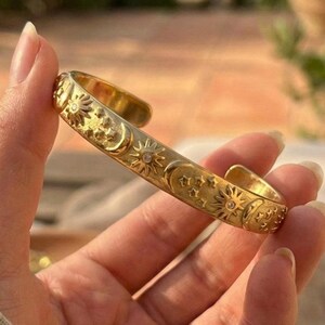 May include: A gold-toned metal cuff bracelet featuring an embossed pattern of suns, moons, and stars. The surface of the bracelet has a polished finish with small, clear, circular accents embedded within the celestial motifs along the band.