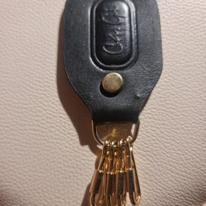 Pode incluir: Um porta-chaves de couro preto com um botão de pressão de metal dourado e quatro ganchos de metal dourado presos a uma argola em D. O componente de couro tem um design oval em relevo com o texto cursivo "Chi Chi" na parte frontal.