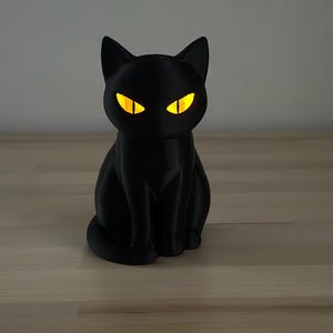 May include: A matte black cat figurine with pointed ears and a seated posture. The cat features glowing, almond-shaped yellow eyes that emit a bright light, creating a sharp contrast against the dark, solid-colored body of the feline sculpture.