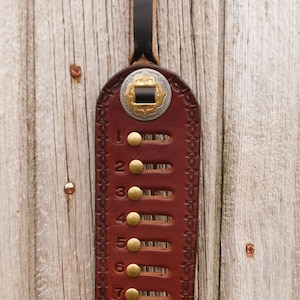 May include: A brown leather horse tack counting tool with brass accents and numbers 1 through 10. The tool is hanging on a wooden wall. The text "Soda Brook Tack" is visible at the bottom of the image.