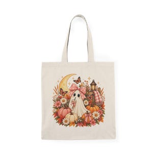 Könnte beinhalten: Eine beigefarbene Stofftasche mit einem zentralen Motiv eines weißen Geistes, der eine rosa Schleife trägt und ein Getränk hält. Der Geist ist umgeben von orangefarbenen und rosa Kürbissen, weißen Gänseblümchen, Herbstblättern, Schmetterlingen und einer Laterne.