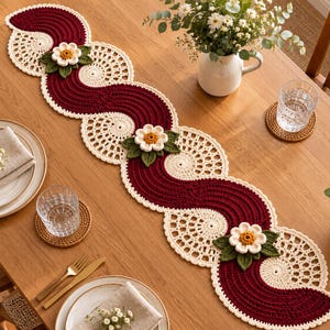 May include: A long, wavy crochet table runner featuring alternating sections of deep burgundy ribbed yarn and cream-colored circular lace patterns. Three dimensional white flowers with yellow centers and dark green leaves are attached.