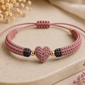 Könnte beinhalten: Ein mauvefarbenes Makramee-Armband mit einem zentralen gehäkelten Herz, zwei kleinen dunkelbraunen gewebten Akzenten und vier polierten goldfarbenen Kugeln. Das verstellbare Kordeldesign hat einen Schiebeknoten und zwei goldene Endperlen.