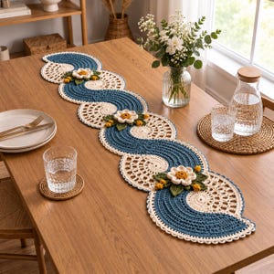 Puede incluir: Un camino de mesa decorativo de ganchillo largo con un patrón de ondas repetitivo en hilo azul verdoso oscuro y crema. El camino de mesa está adornado con motivos florales tridimensionales, incluyendo flores blancas, hojas verdes y pequeños capullos amarillos.