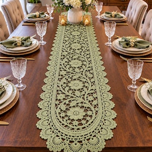 Pode incluir: Um caminho de mesa de renda verde-sálvia com um padrão floral intrincado de motivos circulares e borda festonada, colocado sobre uma mesa de jantar de madeira escura com pratos brancos, guardanapos verdes e copos de cristal transparente.