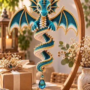 May include: A crocheted dragon ornament in shades of teal, cream, and gold, featuring large wings, a coiled tail, and a faceted blue crystal drop at the bottom. The dragon is suspended from a curved wooden stand, showcasing intricate yarn work.