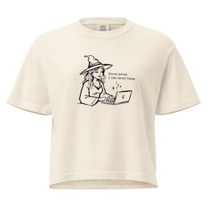 May include: A cream-colored short-sleeve t-shirt featuring a black line-art graphic of a person wearing a witch hat while typing on a laptop. The text "hocus pocus I can never focus" is printed next to the figure, along with small star icons.